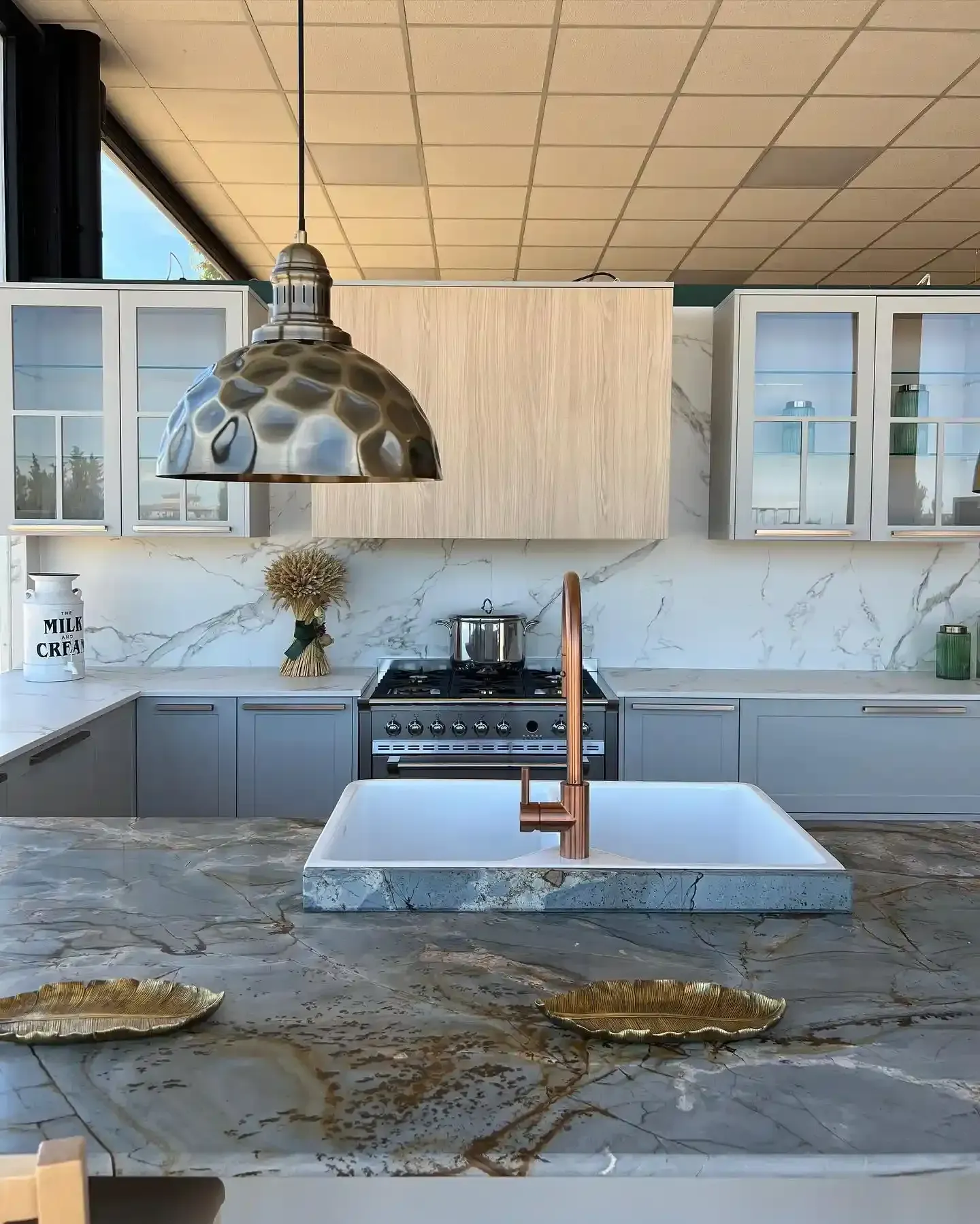 Cocina moderna con isla de mármol, fregadero estilo rústico, lámpara colgante y armarios grises.