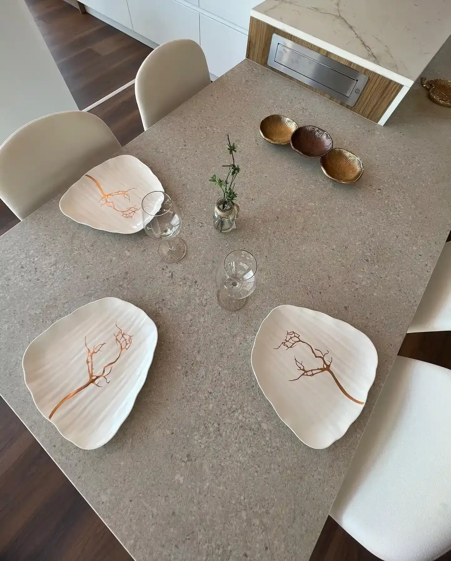 Mesa de comedor puesta con platos blancos, un pequeño jarrón central y tres cuencos decorativos sobre un tablero gris.