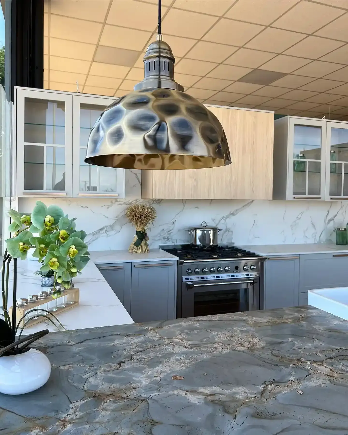 Cocina moderna con gabinetes grises, salpicadero de mármol, isla de piedra y una gran lámpara colgante con forma de cúpula.