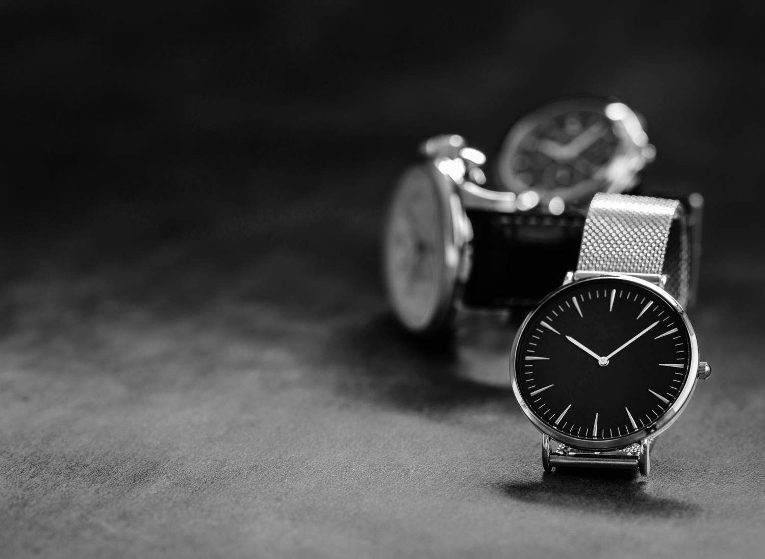 Imagen en blanco y negro: Primer plano de un reloj de pulsera plateado con esfera negra, otros relojes borrosos.
