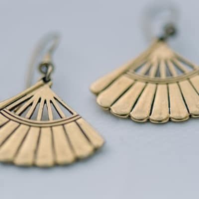 Pendientes dorados en forma de abanico con un diseño texturizado y detallado, colgando sobre un fondo borroso.