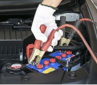 Manos con guantes conectando cables de arranque rojos a la batería de un coche azul en el compartimento del motor