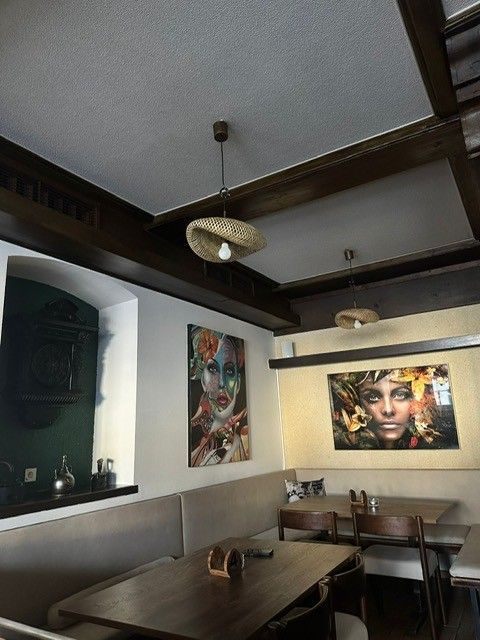 Eine Restaurantecke mit Holztischen, beigefarbenen Sitznischen, hängenden Lichterketten und zwei großen künstlerischen Porträts an den Wänden.