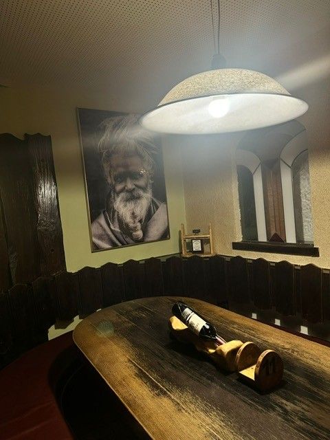 Ein rustikaler Holztisch mit einer Weinflasche im Halter, daneben ein Wandporträt und eine Pendelleuchte in einem schwach beleuchteten Raum.