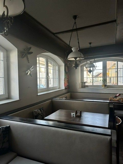 Ein leeres Restaurantinterieur mit plüschigen beigefarbenen Sitznischen, Holztischen und Hängelampen in der Nähe von Bogenfenstern.