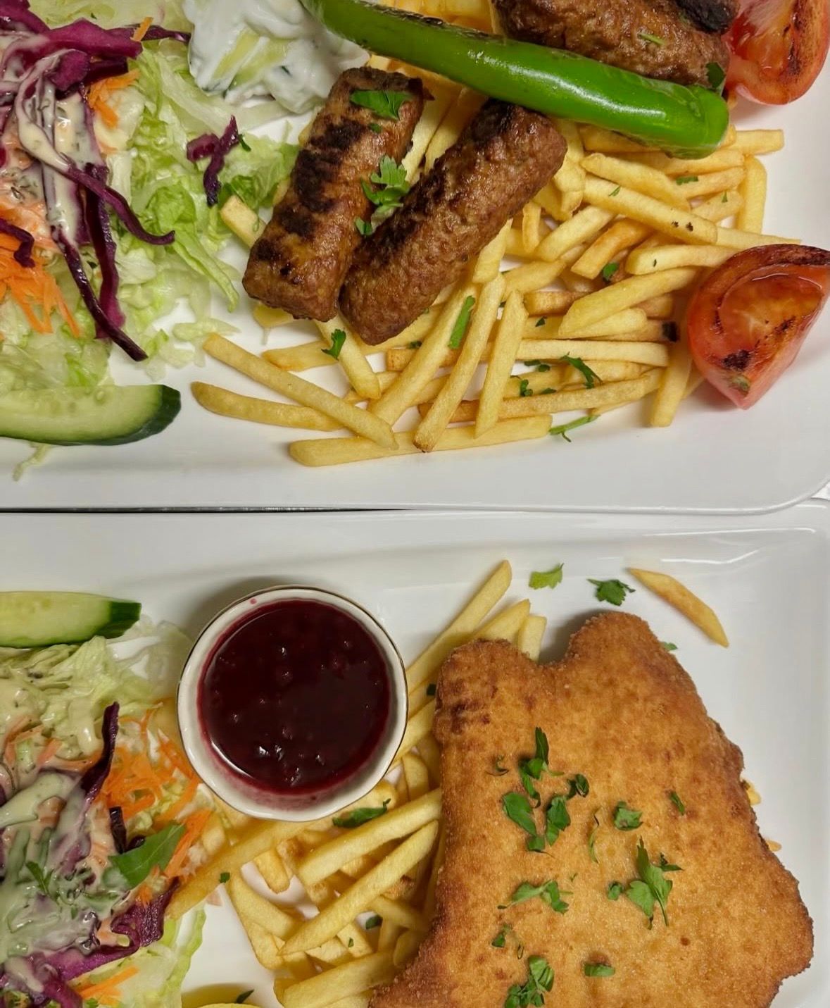 Zwei Teller mit Essen, einer mit gegrillten Würstchen, Pommes frites und Salat, der andere mit einem panierten Schnitzel mit Soße.