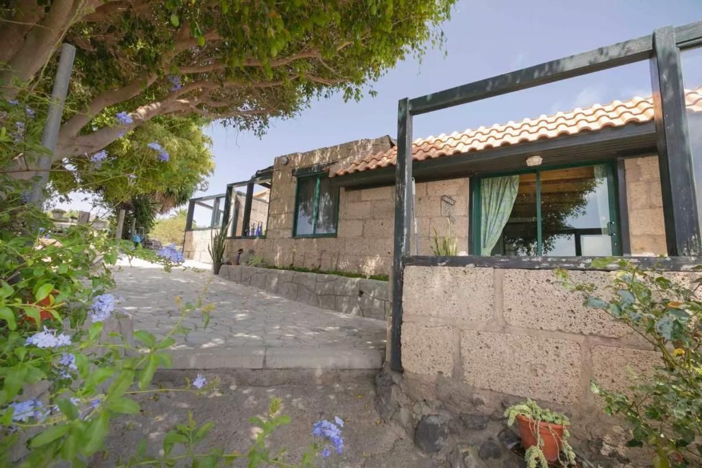 Cabaña de piedra con techo de tejas y estructura de madera al aire libre. Un sendero pavimentado conduce a la entrada; flores y árboles flanquean la construcción.