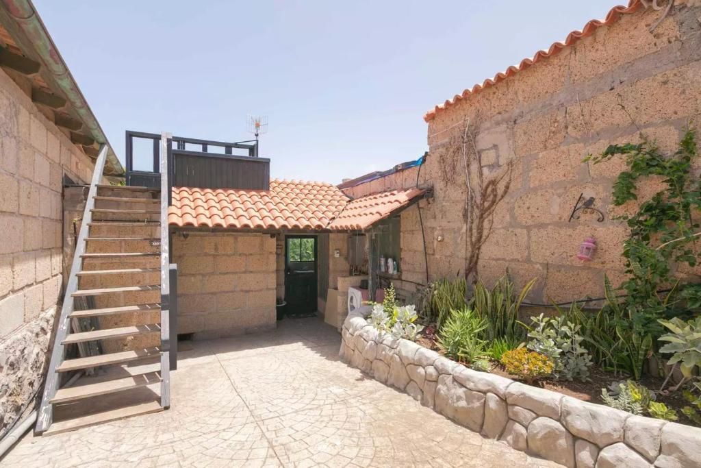 Patio con edificios de piedra, techo de terracota, escaleras a una terraza en la azotea y un jardín con plantas.