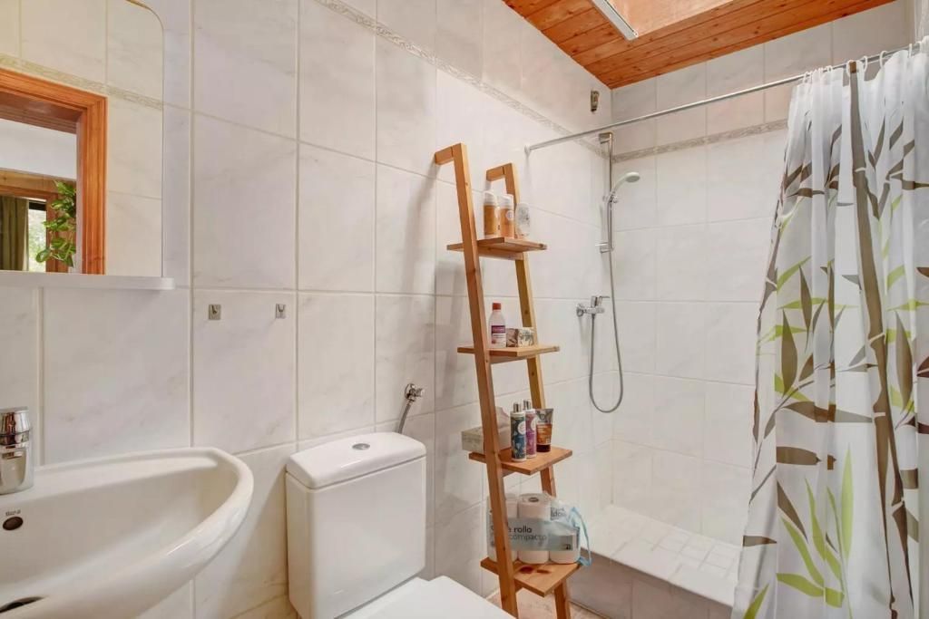 Un baño pequeño con paredes de azulejos blancos, inodoro, lavabo y ducha. Una escalera de madera contiene artículos de aseo y una cortina de ducha con estampado de bambú.