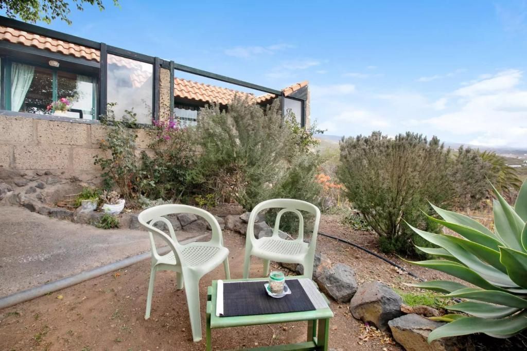 Dos sillas de plástico blancas y una mesa pequeña se encuentran en un patio con vistas a un paisaje, con un edificio al fondo.