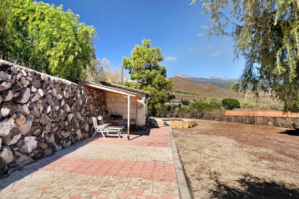 Patio con muro de piedra, pavimento de ladrillo rojo y gris, y una pequeña estructura con mesa y sillas. Al fondo se ven árboles y una cordillera.