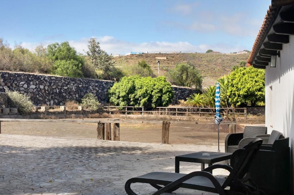 Una escena soleada al aire libre con sillas en un patio, con vista a un campo seco, un muro de piedra y vegetación.