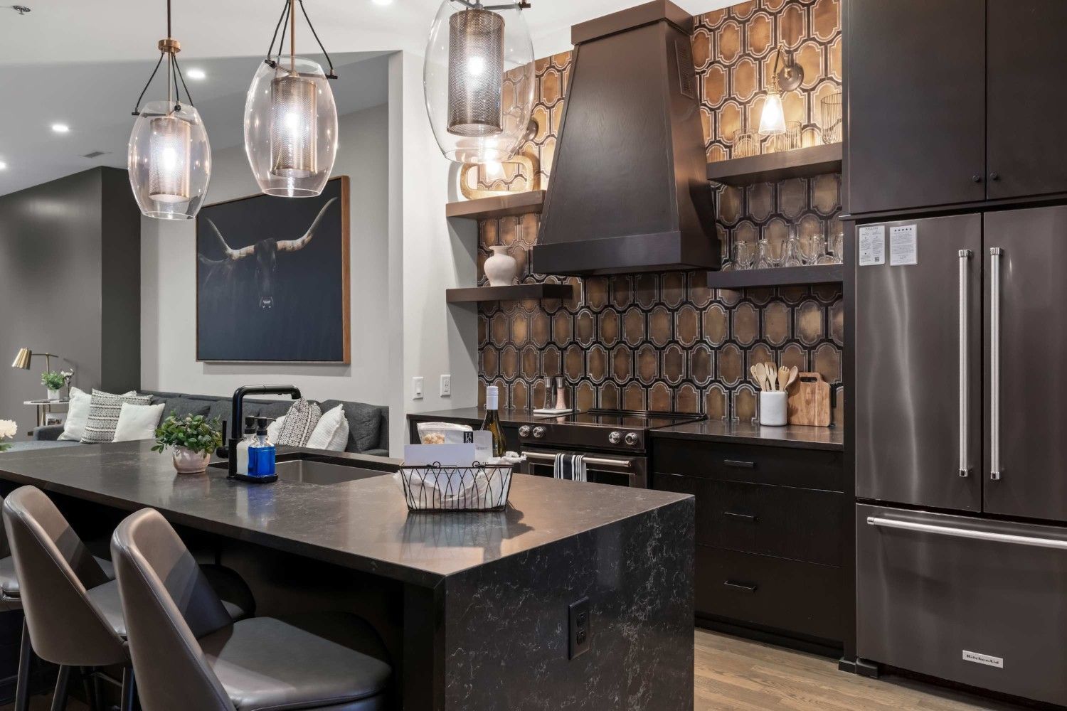 A modern kitchen with dark cabinetry, a long island with seating, a large range hood, and gold hexagonal backsplash tiles.