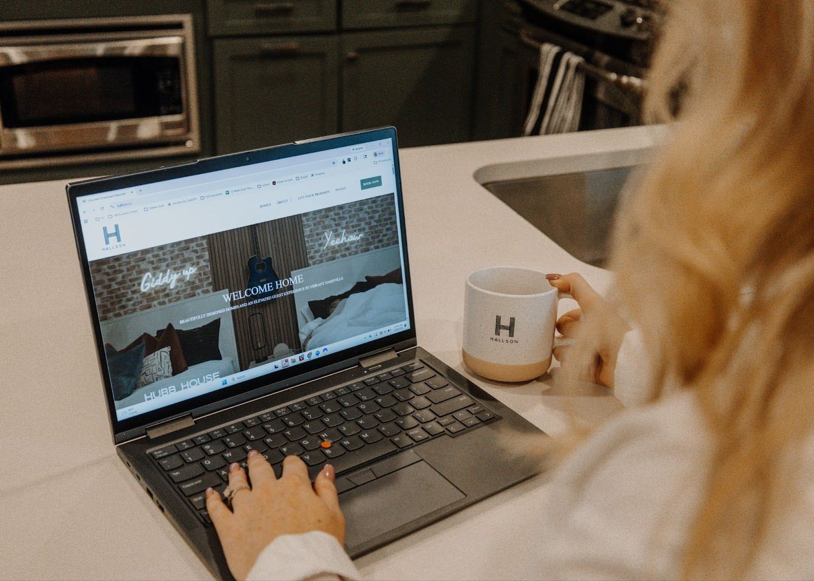 A person types on a laptop in a kitchen, with a branded mug beside the computer on a white countertop.