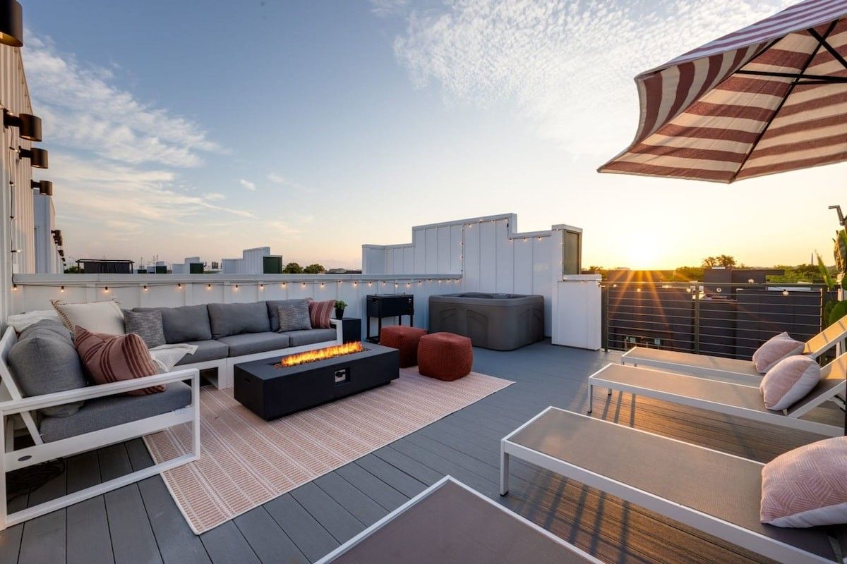 A rooftop patio at sunset featuring a gray sectional sofa, a fire pit, lounge chairs, and an umbrella on a wooden deck.