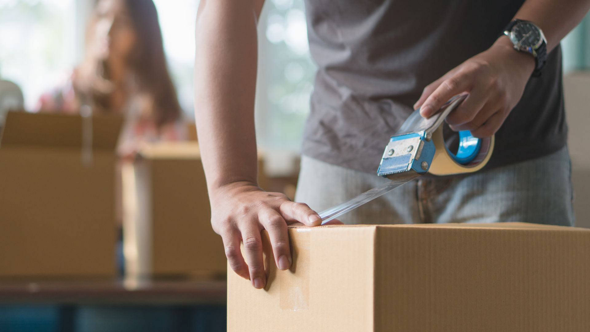 Une personne ferme une boîte en carton avec du ruban adhésif. D'autres boîtes sont visibles en arrière-plan.