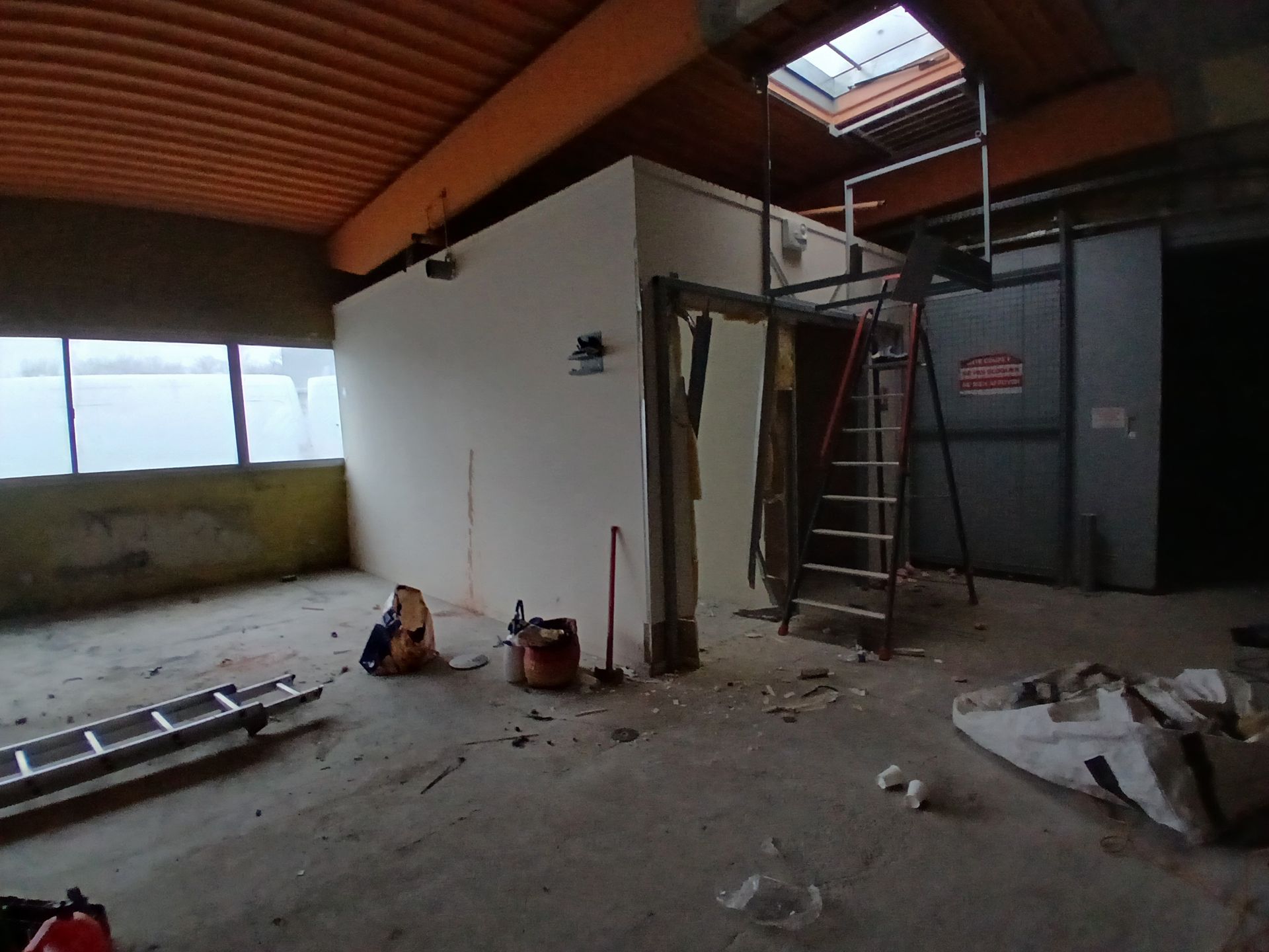 Chantier de construction intérieur avec un mur partiellement démoli, une échelle et des débris sur un sol en béton. Ceci est avant l'intervention du professionnel.