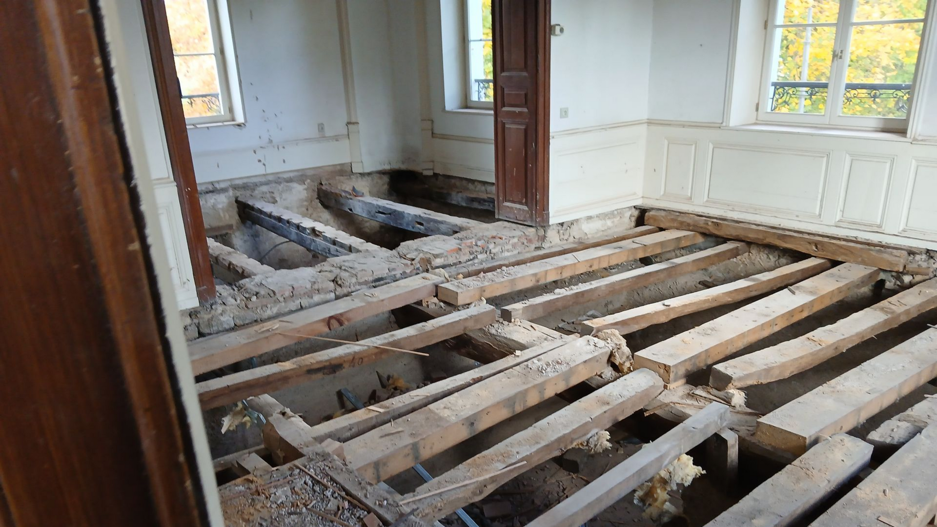 Pièce avec solives apparentes, révélant un sous-plancher retiré. On aperçoit des fenêtres et une porte.
