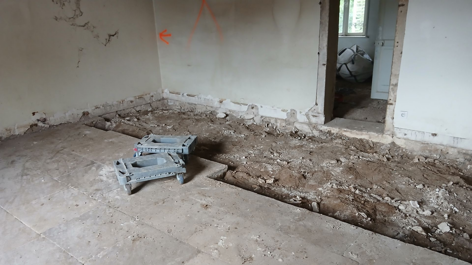 Pièce en travaux, avec sol apparent et gravats. Murs et porte gris.