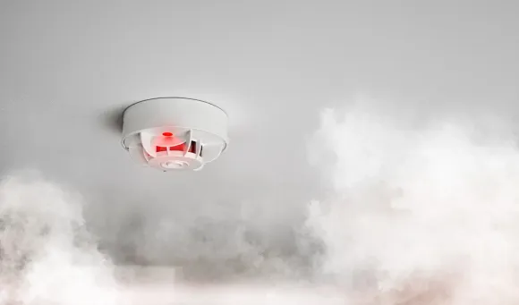 Detector de humo en el techo con luz roja intermitente, rodeado de humo.