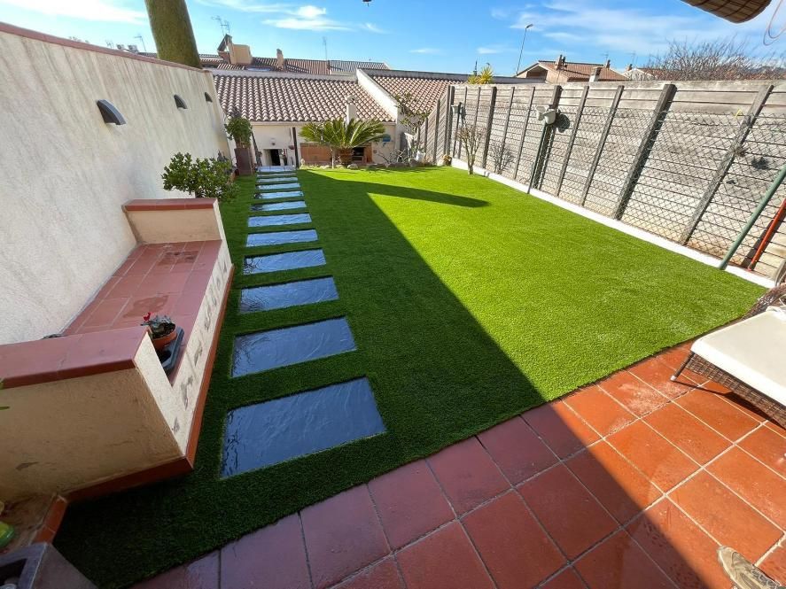 Un patio trasero con césped artificial verde, un sendero de losas de piedra, un banco bajo de color beige