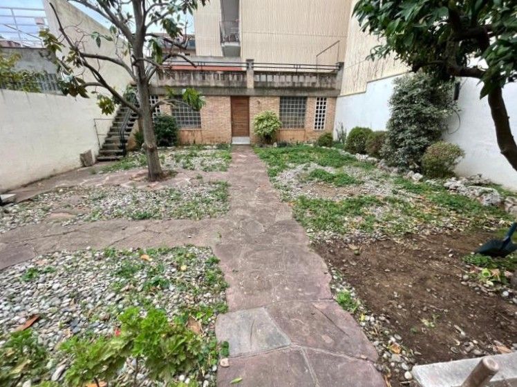 Un sendero de piedra atraviesa un jardín con parterres de grava, plantas bajas y árboles, y conduce a un edificio de ladrillo
