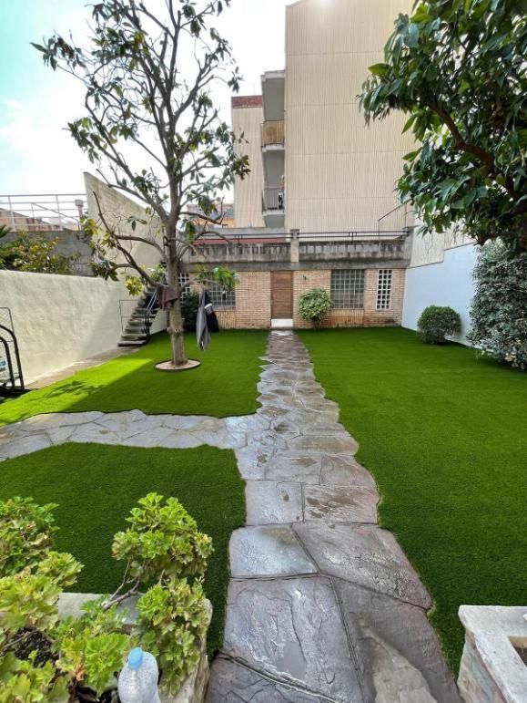 Un sendero de piedra atraviesa un césped artificiae en un patio, flanqueado por árboles y un edificio de piedra.