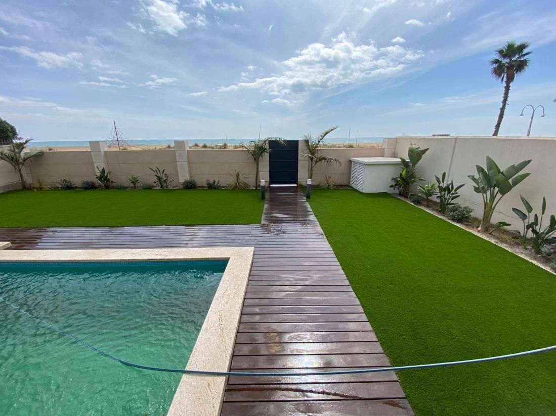 Una piscina con un sendero de madera que conduce a una puerta en un muro, con vistas al océano bajo un cielo azul.