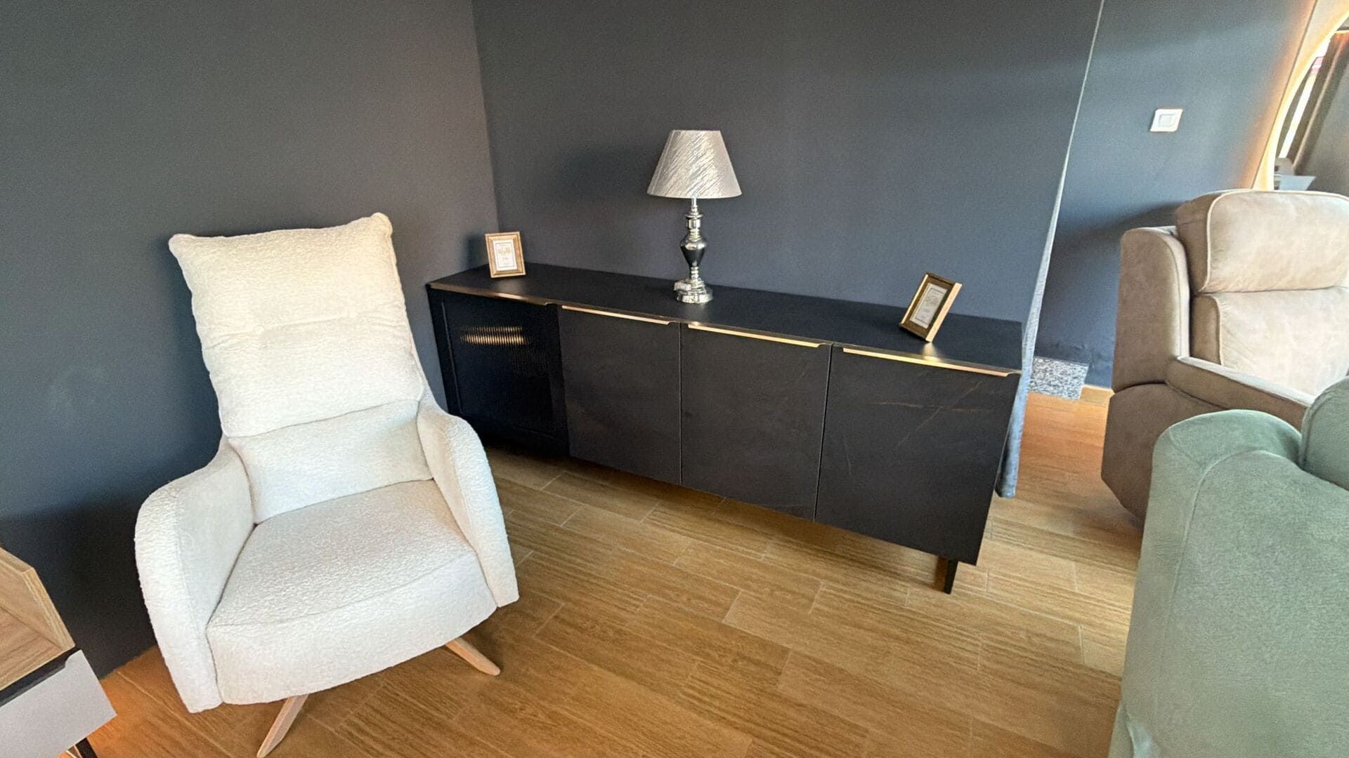 Salón con armario negro, sillón color crema, lámpara, detalles dorados y suelo de madera.