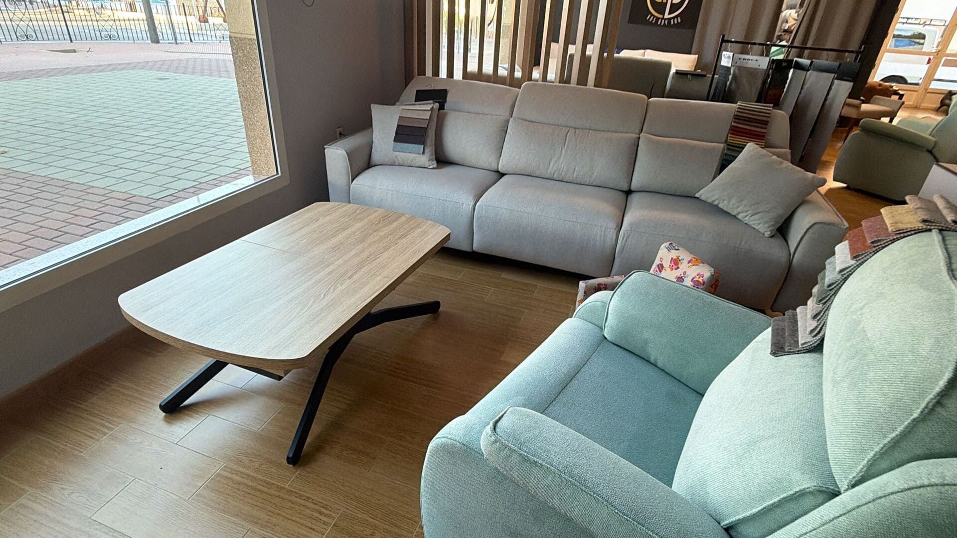 Sala de estar con sofá de color claro, sillón azul verdoso, mesa de centro de madera y gran ventanal con azulejos de mosaico.