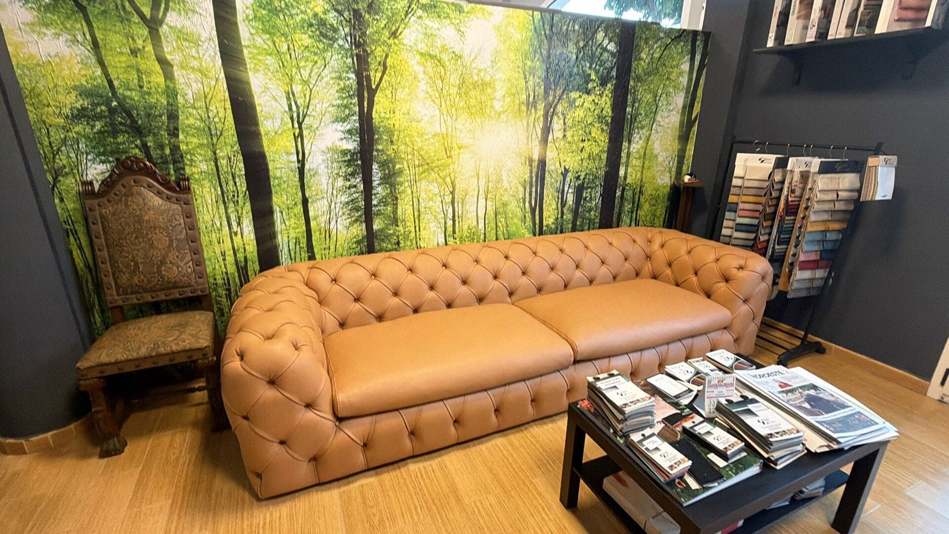 Sofá de cuero capitoné color canela en una habitación con papel pintado de bosque, silla antigua y revistas sobre una mesa.