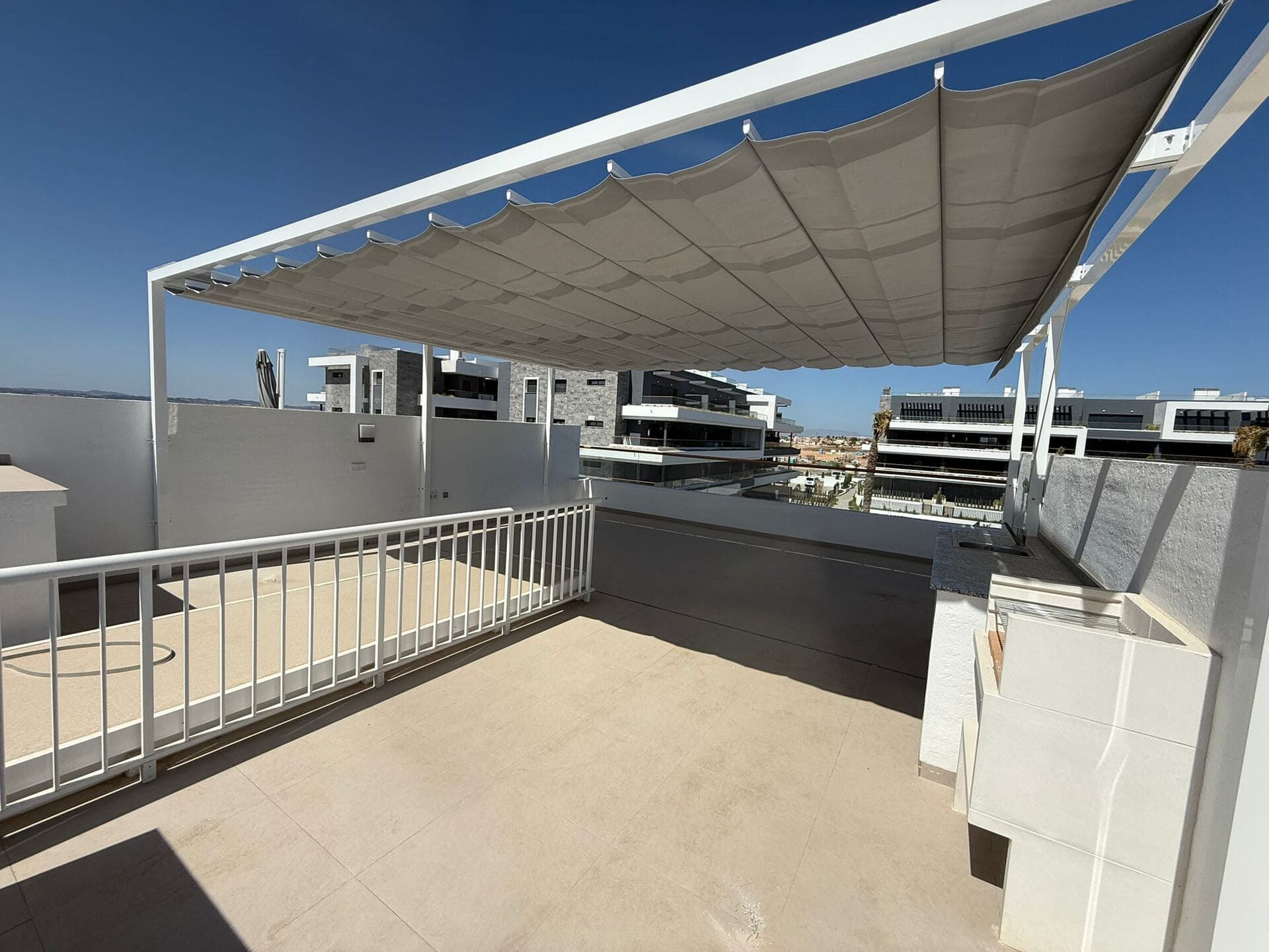 Terraza en la azotea con barandilla blanca, toldo y edificios en construcción al fondo. Día soleado.