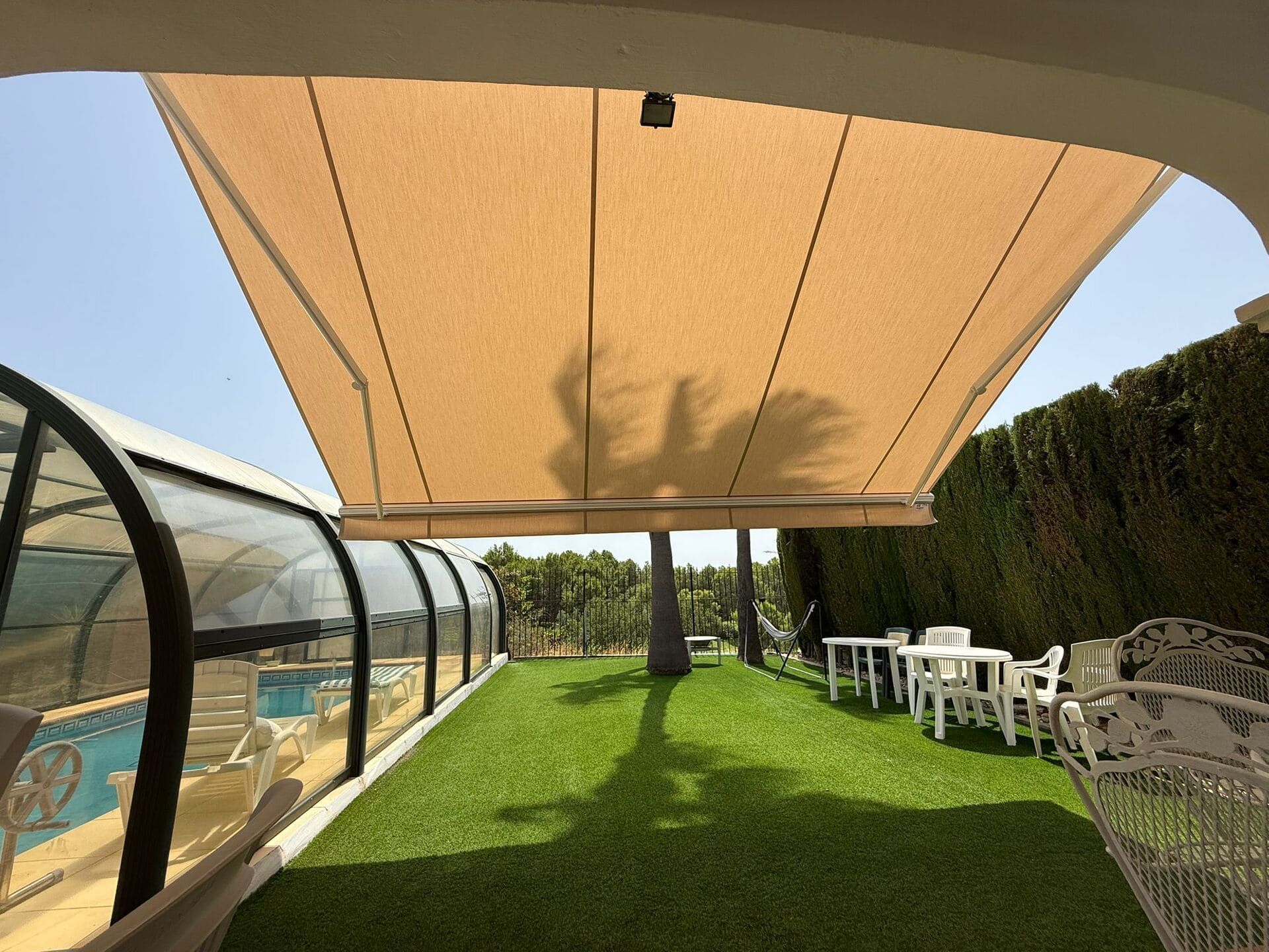 Toldo color beige sobre un césped verde con una palmera, cerramiento de piscina y muebles de exterior blancos.