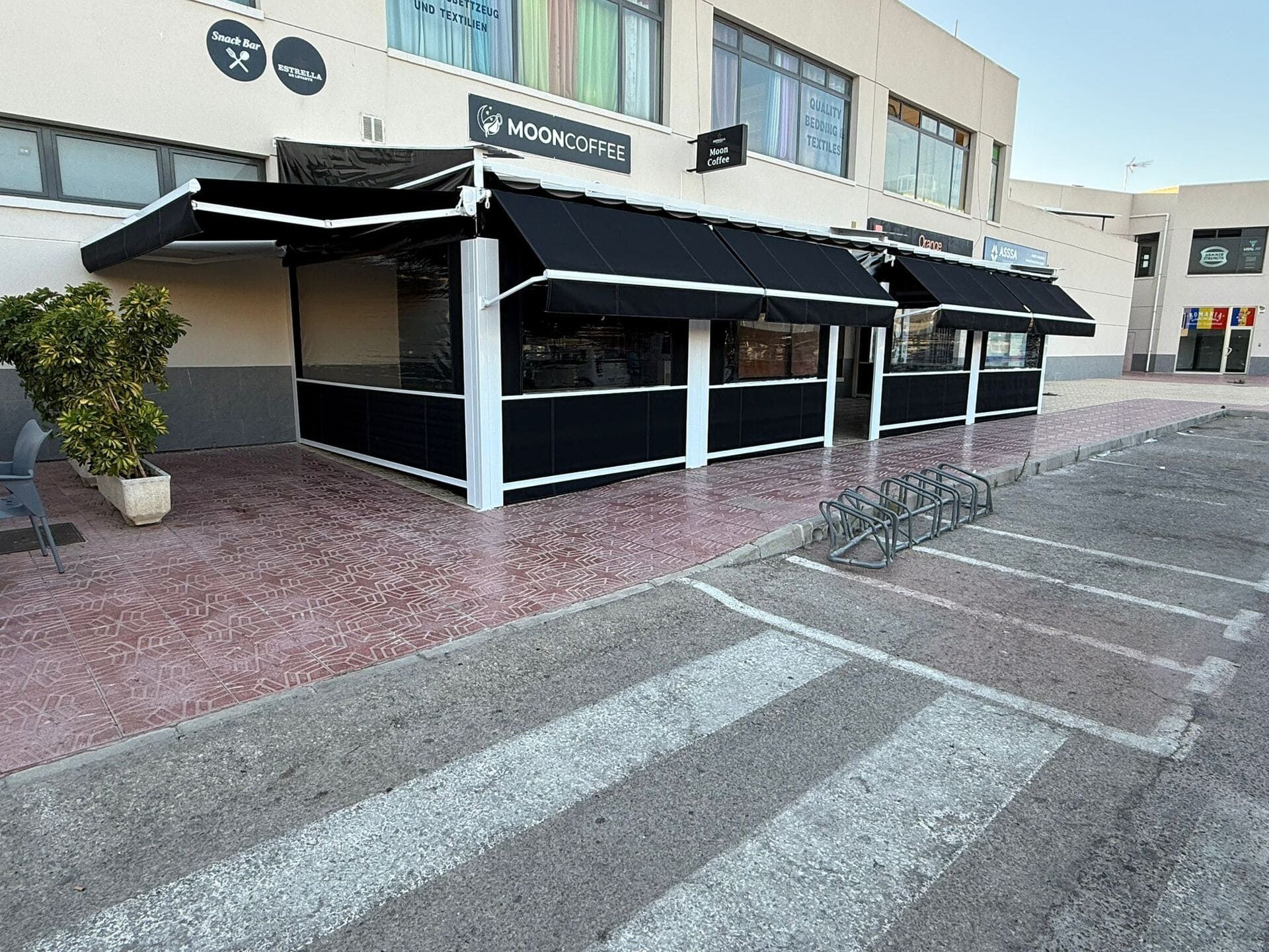 Exterior de un negocio con toldos negros, molduras blancas y acera de color ladrillo, aparcamiento para bicicletas, edificio.
