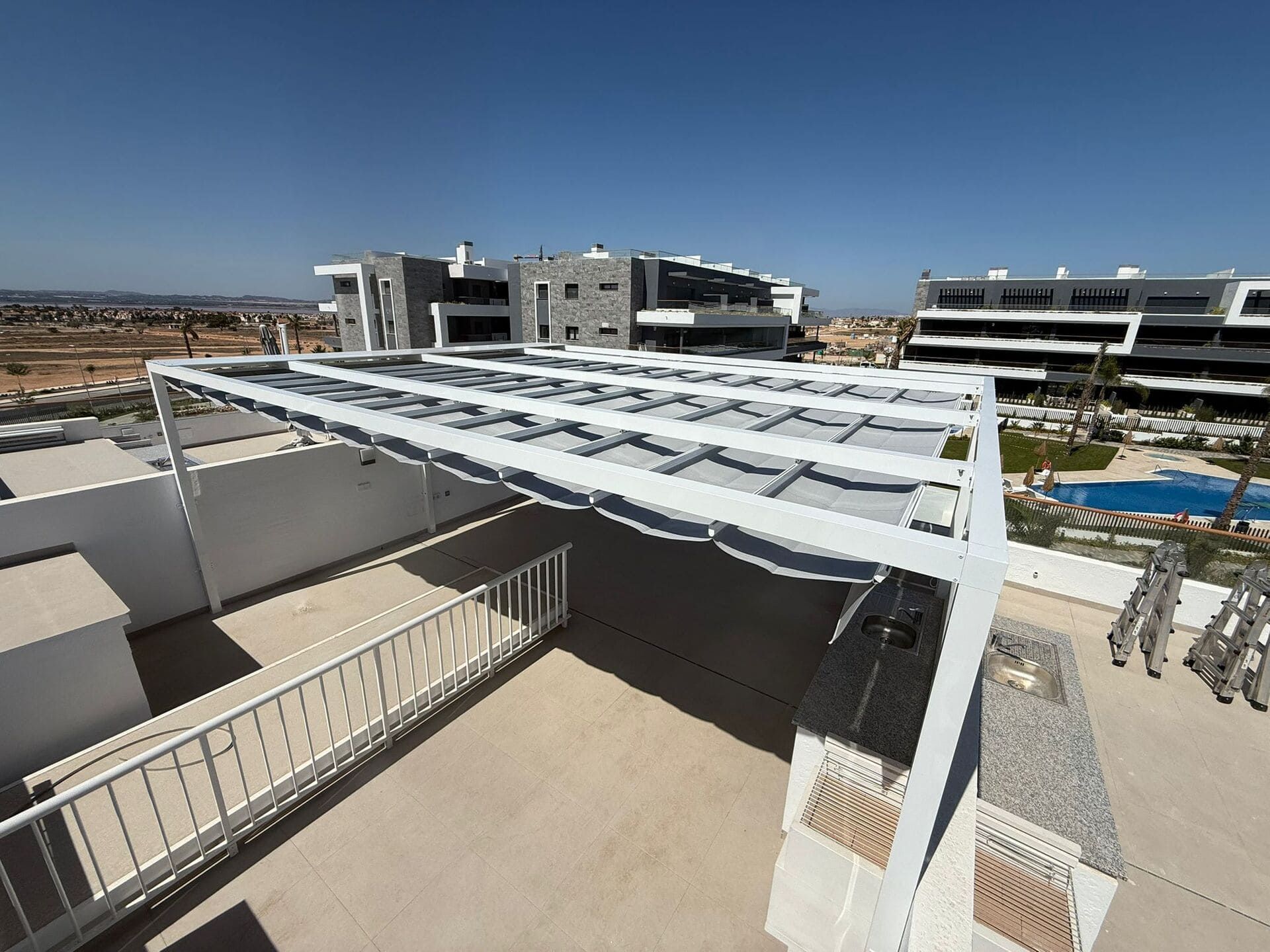 Terraza en la azotea con pérgola blanca que da sombra. Edificios modernos y piscina al fondo, bajo un cielo azul.