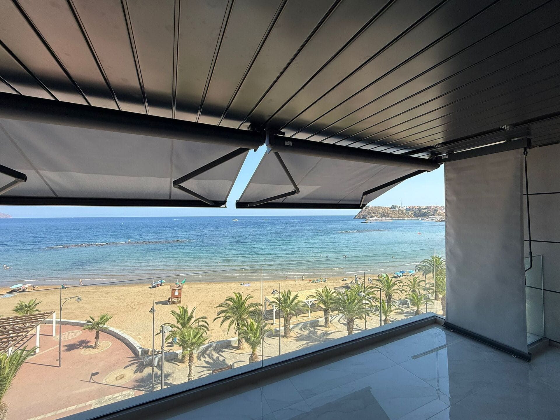Vista de la playa y el océano desde debajo de un toldo retráctil. Palmeras, cielo azul y una orilla arenosa.