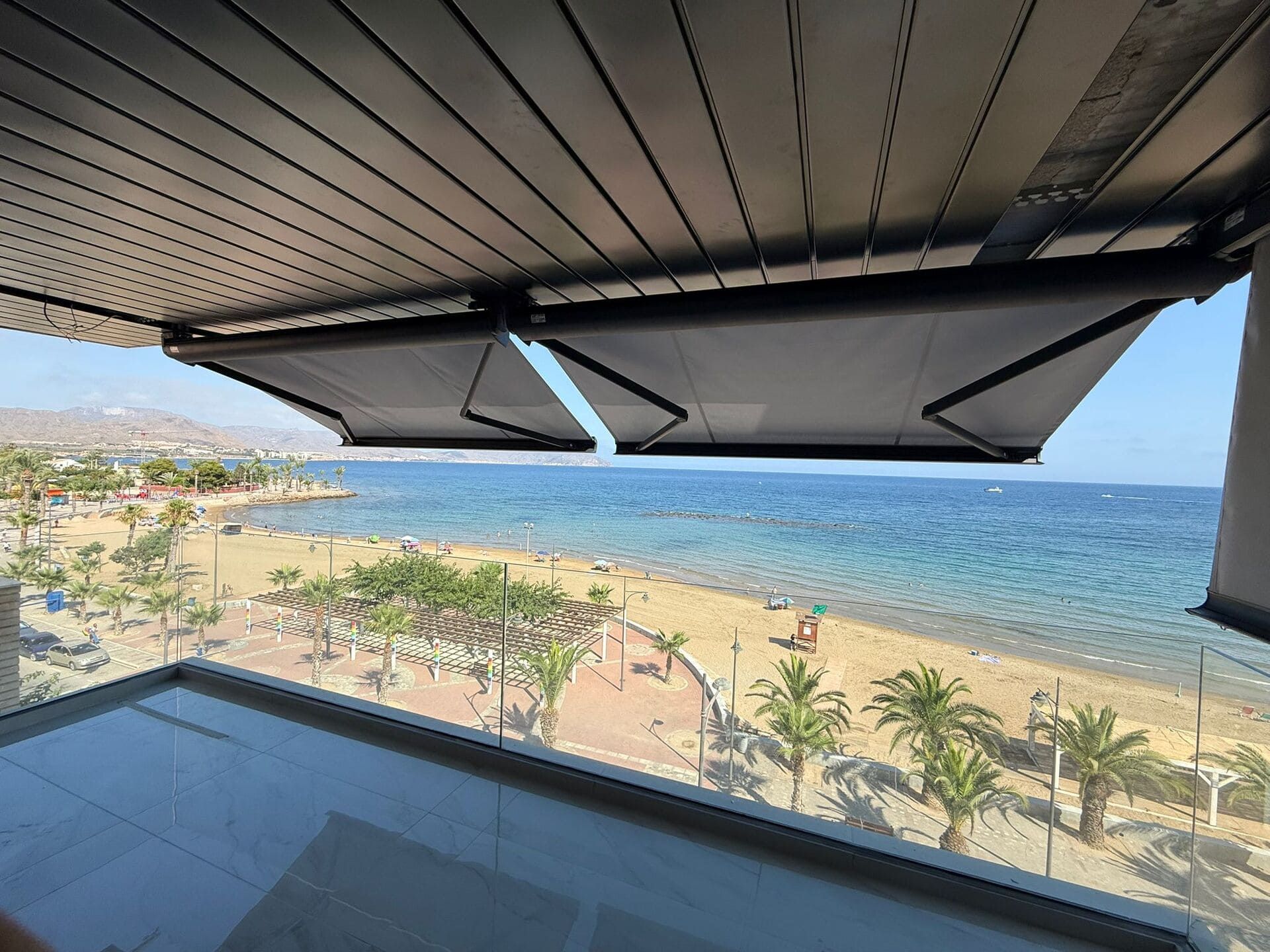 Dos toldos retráctiles dan sombra a un balcón con vistas a una playa con palmeras y al océano.