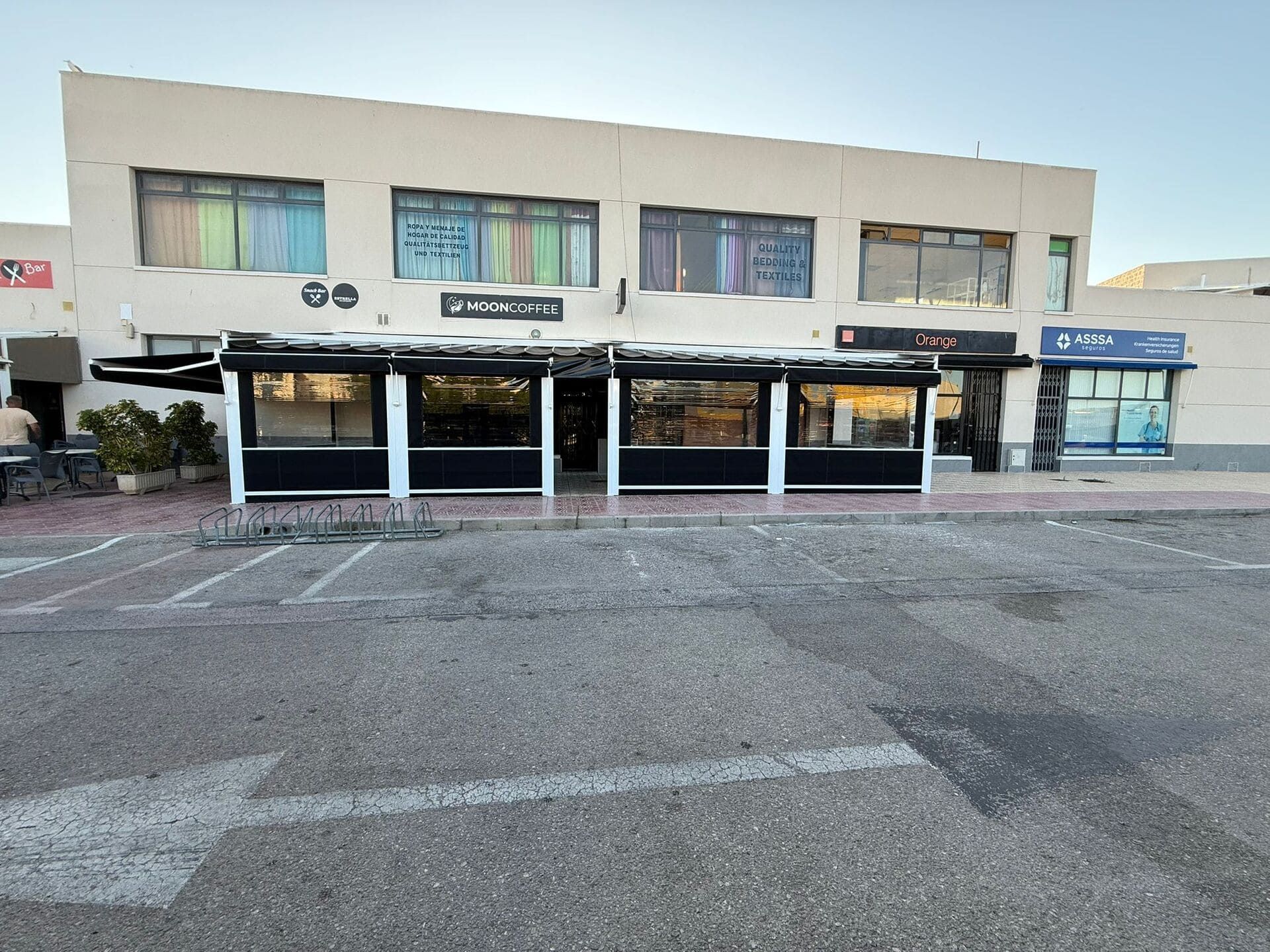 Exterior de un edificio de dos plantas con tiendas y terraza en un aparcamiento bajo un cielo despejado.