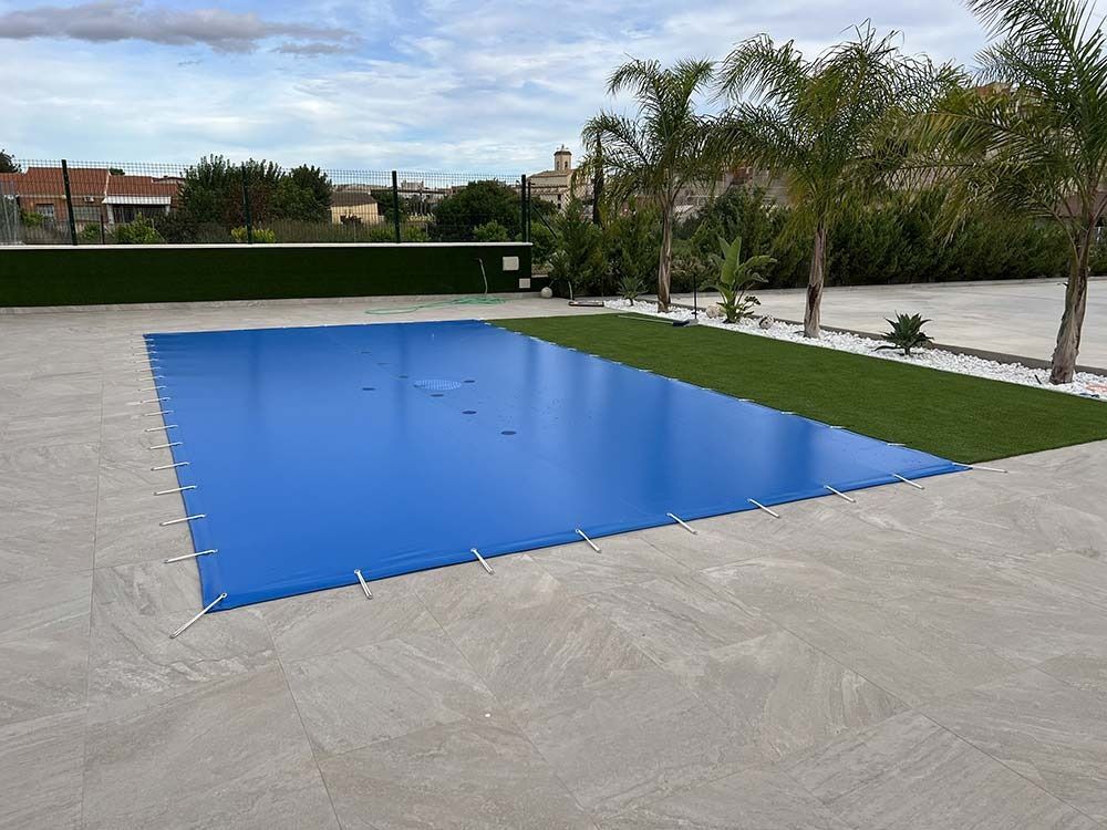 Lona azul sobre una piscina rectangular. Patio de hormigón, césped verde, palmeras al fondo.