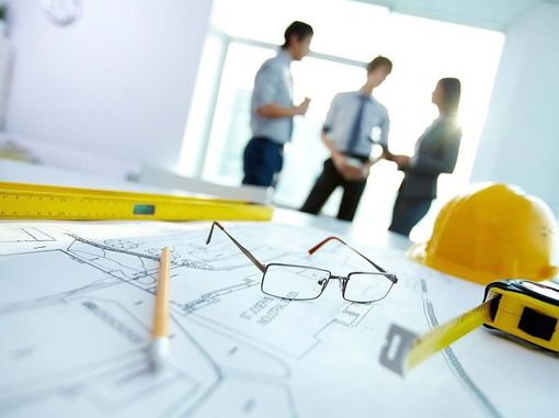 Planos arquitectónicos con herramientas (gafas, nivel, cinta métrica, casco) sobre una mesa, tres personas borrosas al fondo.