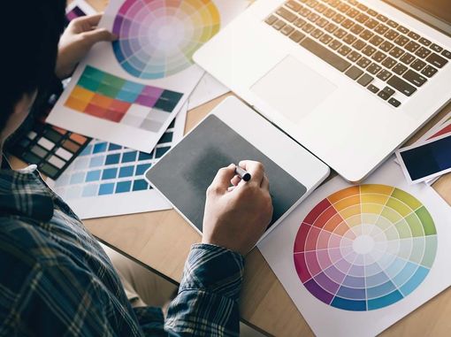 Persona diseñando en una tableta, rodeada de paletas de colores y un ordenador portátil.