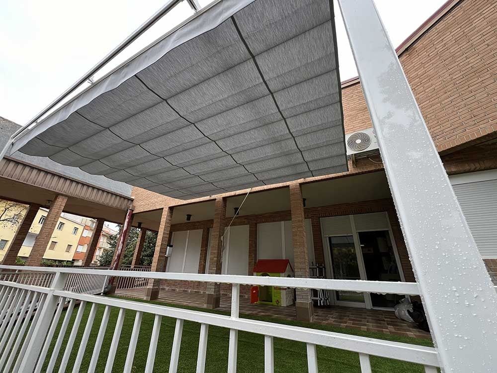 Un toldo gris para dar sombra sobre un patio, con soportes metálicos blancos y una valla blanca.