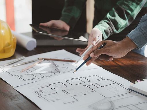 Dos personas revisando planos arquitectónicos en un escritorio con una computadora portátil, un lápiz y un casco.