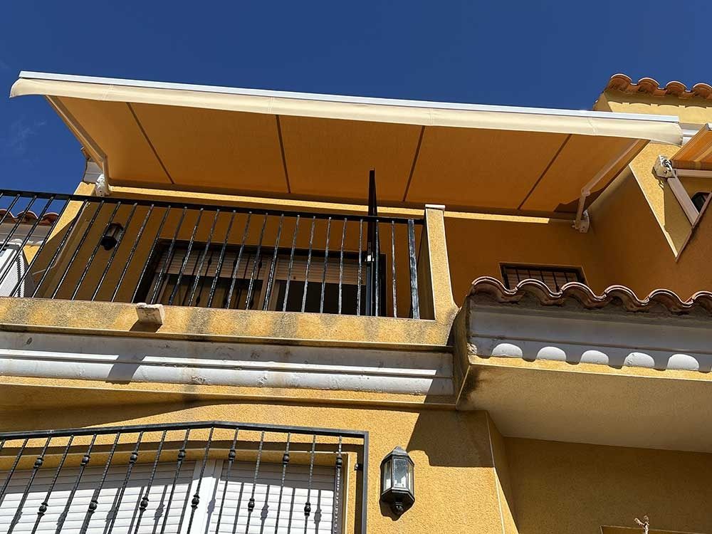 Edificio amarillo con balcón y toldo; barandilla negra.
