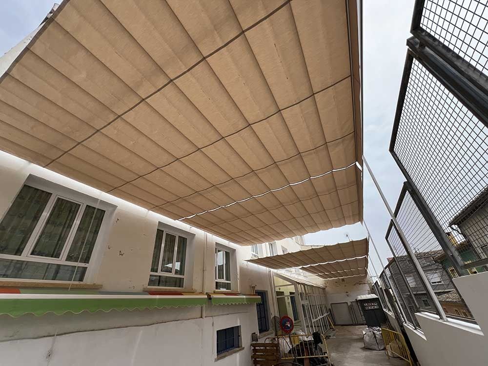 Un toldo color beige da sombra al espacio al aire libre de un edificio, con ventanas y una valla de tela metálica a la derecha.