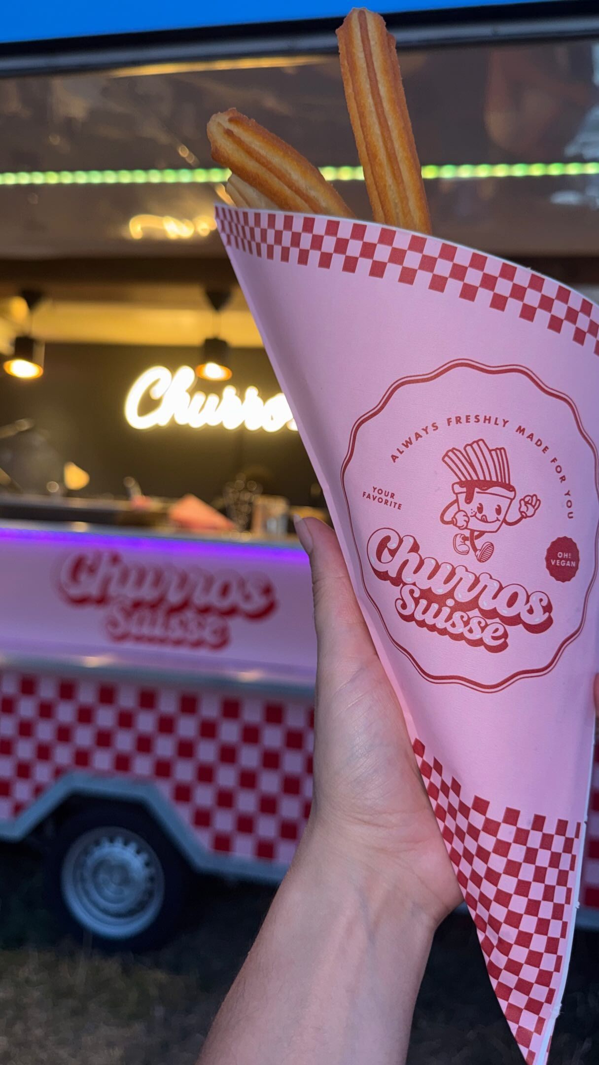 Eine Hand hält eine rosa karierte Waffel an einem Craveous-Foodtruck vor warmem Licht.