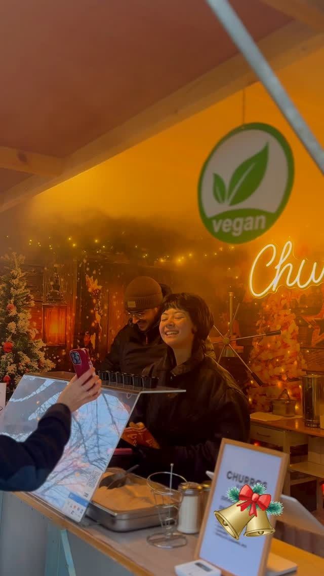 Frau lächelt hinter einem veganen Marktstand mit warmem Licht und einem Weihnachtsschild