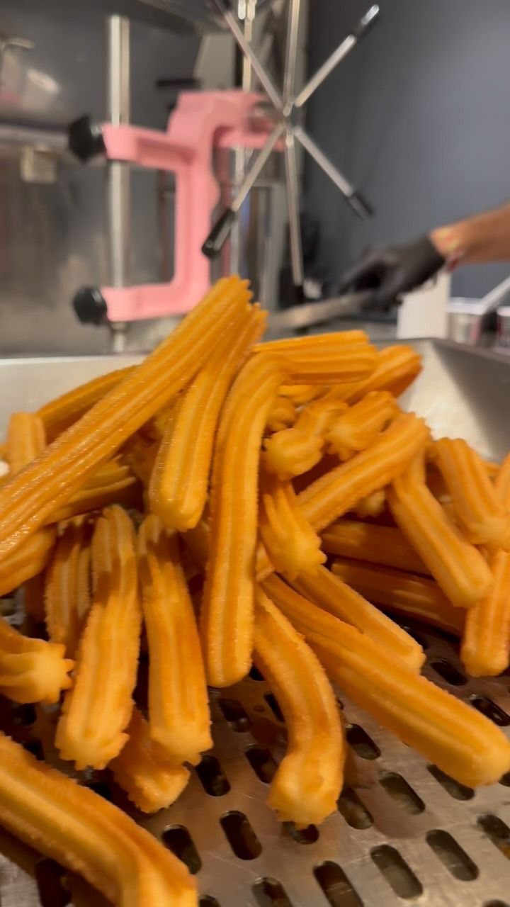 Goldene Churros auf einem Metalltablett gestapelt, im Hintergrund verschwommen eine rosafarbene Maschine.