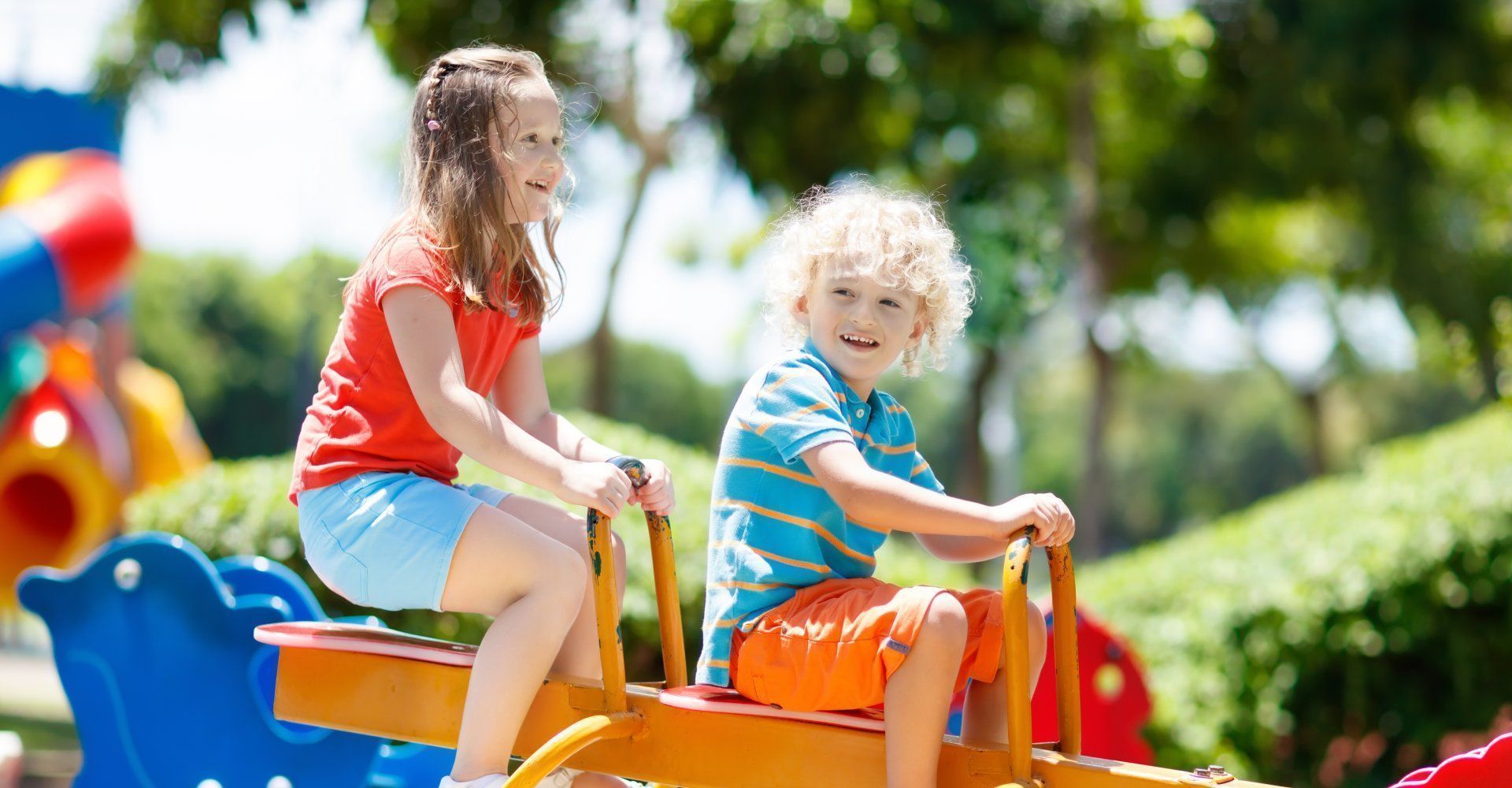 Zwei Kinder auf einer Wippe auf einem bunten Spielplatz, lächelnd, Bäume im Hintergrund.