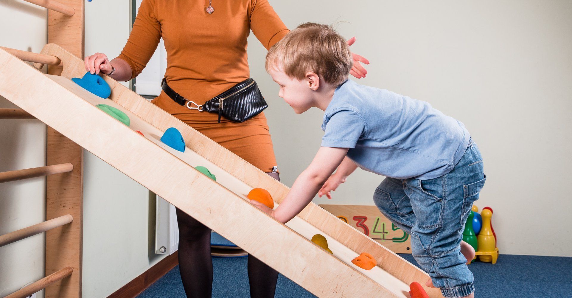 Ein kleiner Junge klettert in einer Therapieumgebung unter Anleitung eines Erwachsenen an einer bunten Kletterwand aus Holz hoch.